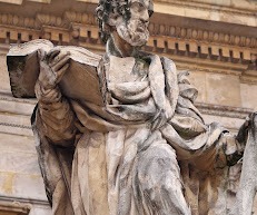 Kamienna rzeźba mężczyzny z brodą, trzymającego otwartą księgę na tle zdobionej fasady budynku.