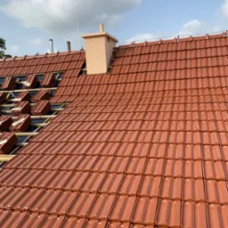 Częściowo pokryty dach z czerwonej dachówki ceramicznej, widoczna więźba dachowa i komin w tle.