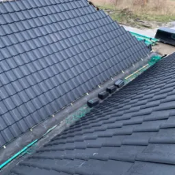 Układanie ciemnej dachówki na dachu domu, widoczna membrana dachowa i zabezpieczenia krawędzi, w tle krajobraz z polami i drzewami.