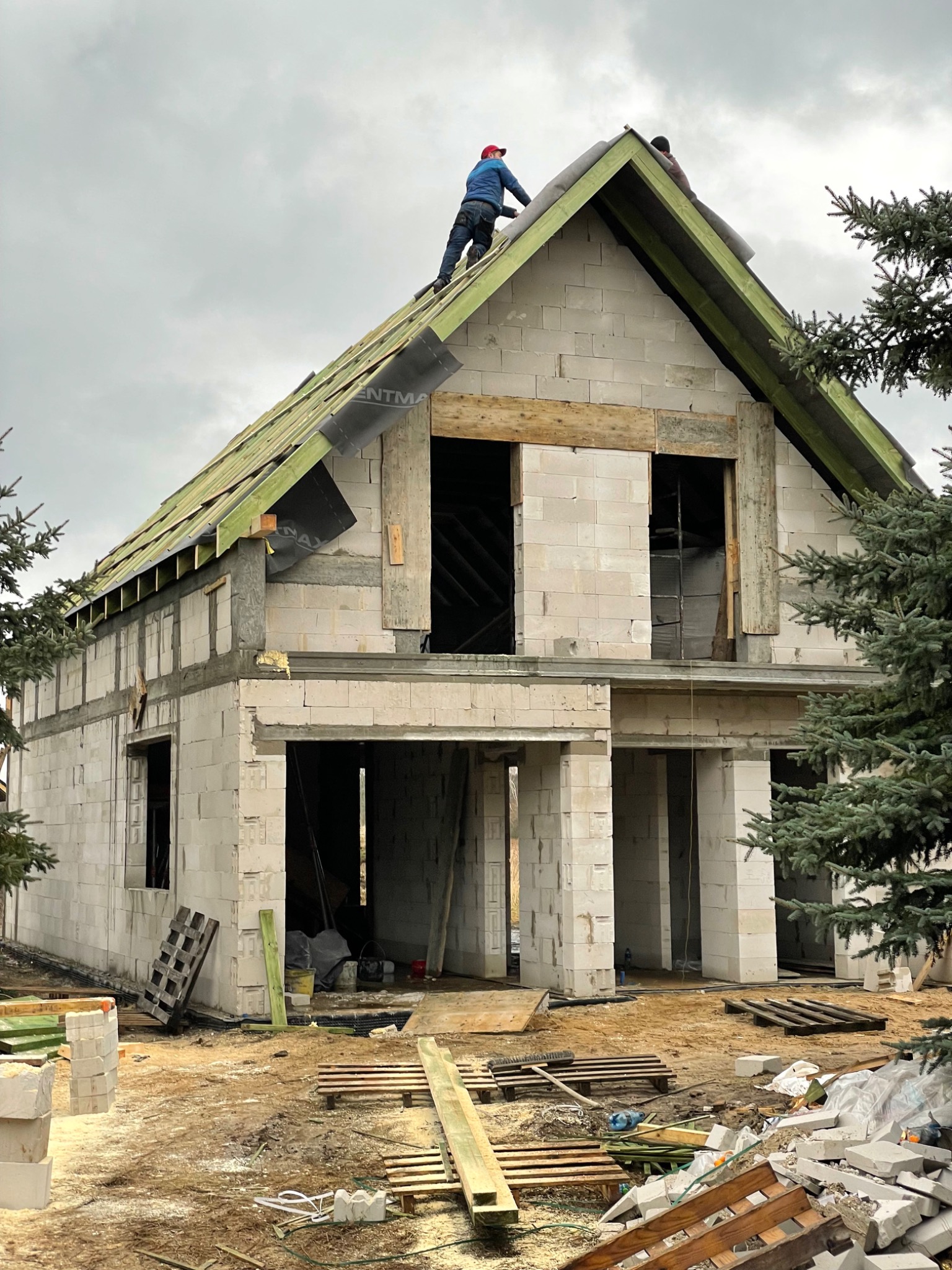 Budowa domu jednorodzinnego z białych bloczków: dwóch robotników na dachu, montaż drewnianej konstrukcji pod pokrycie dachowe, widoczna membrana dachowa i elementy więźby dachowej, teren budowy...