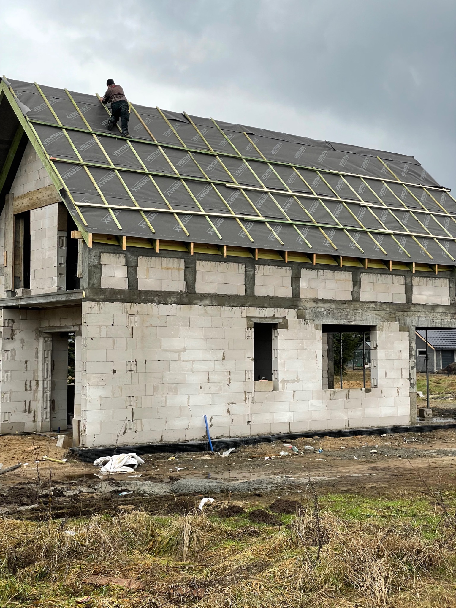 Prace na dachu domu w trakcie budowy: dekarz montuje membranę dachową i łaty pod pokrycie dachowe, widoczne bloczki betonu komórkowego ścian zewnętrznych.