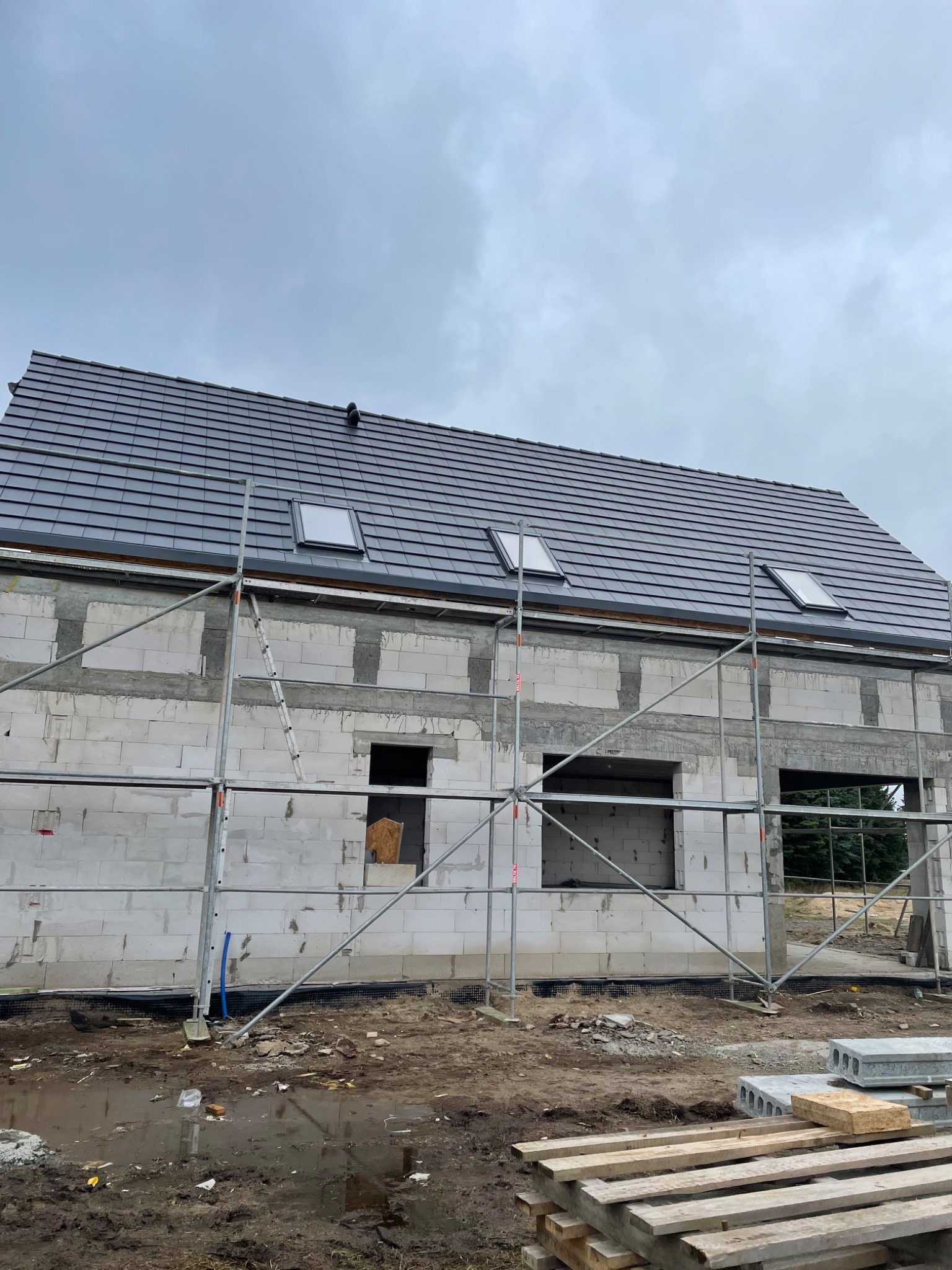 Dom w trakcie budowy z zamontowanym szarym dachem z trzema oknami dachowymi, widoczne rusztowania i teren budowy.