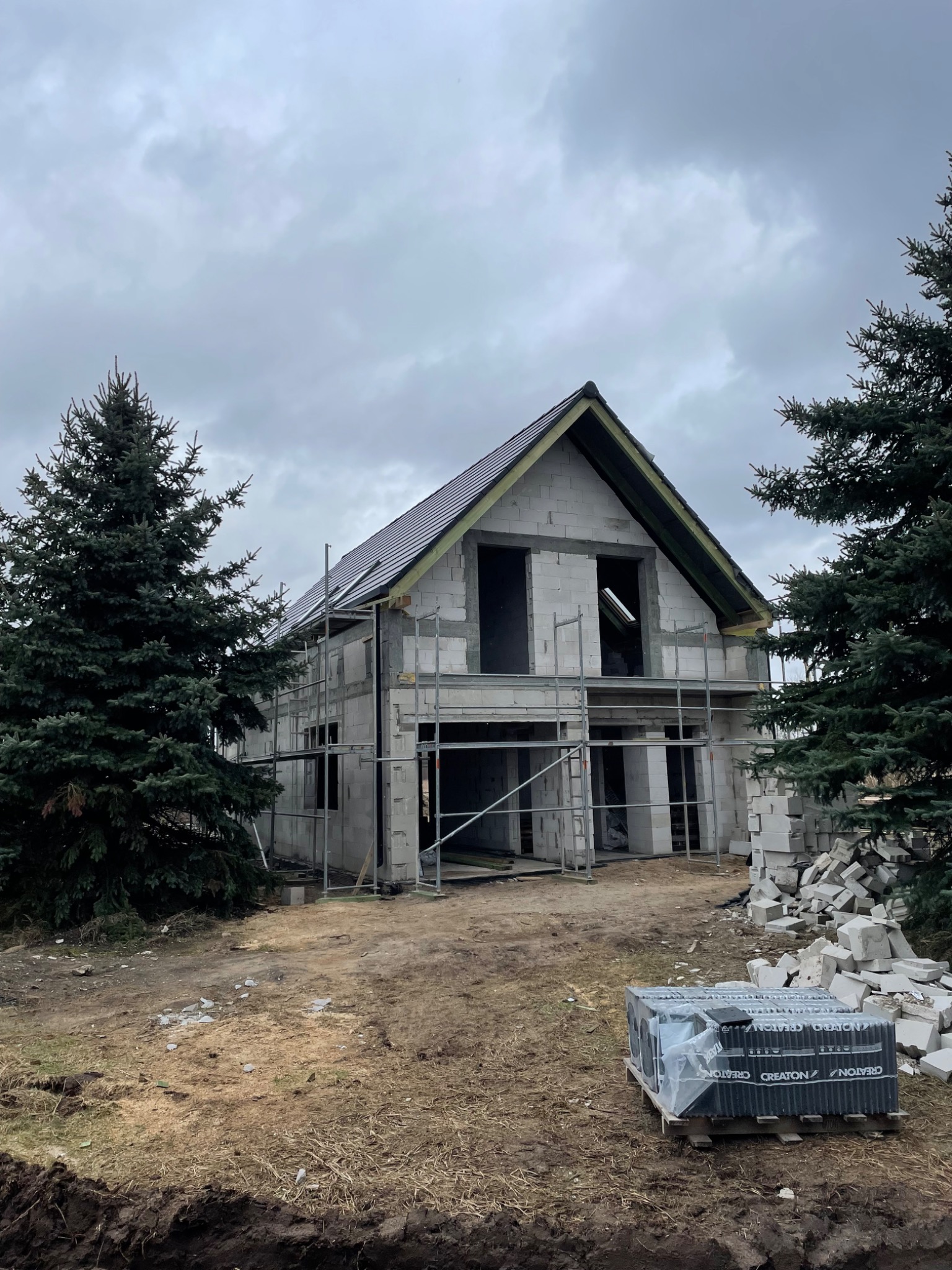 Dom w stanie surowym otwartym z częściowo położonym ciemnym pokryciem, widoczne rusztowania, obok stos bloczków i paleta z dachówkami CREATON, pochmurne niebo.