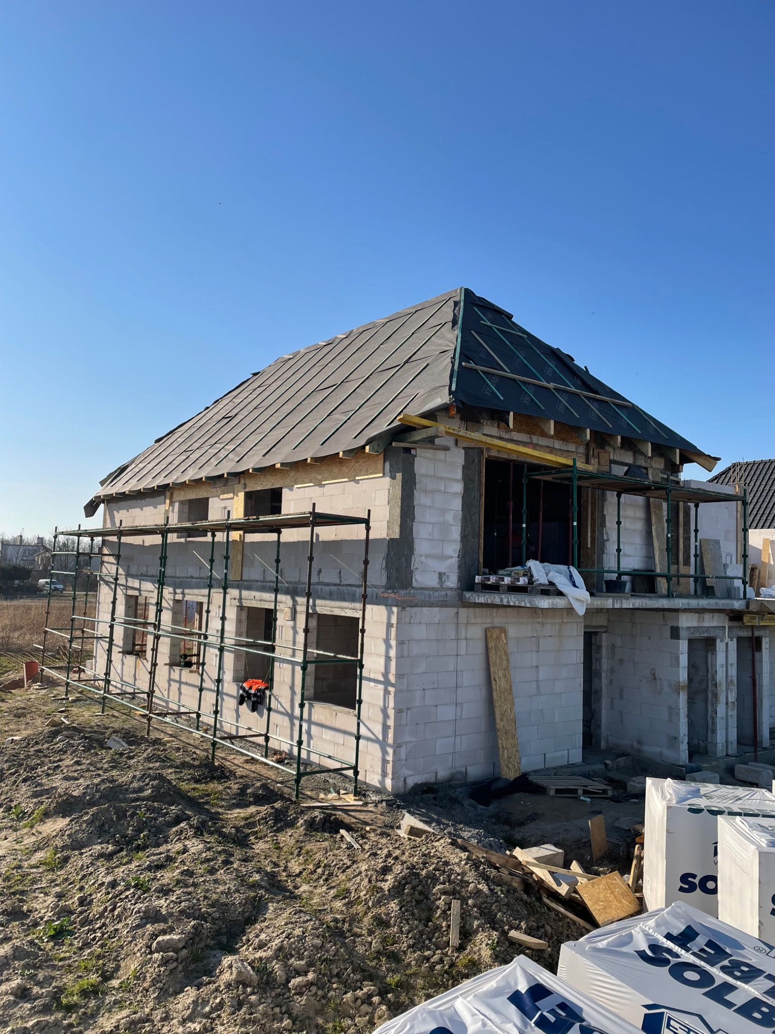Dwupiętrowy dom w trakcie budowy z częściowo położonym pokryciem z papy na dachu, otoczony rusztowaniami i stertami materiałów budowlanych.