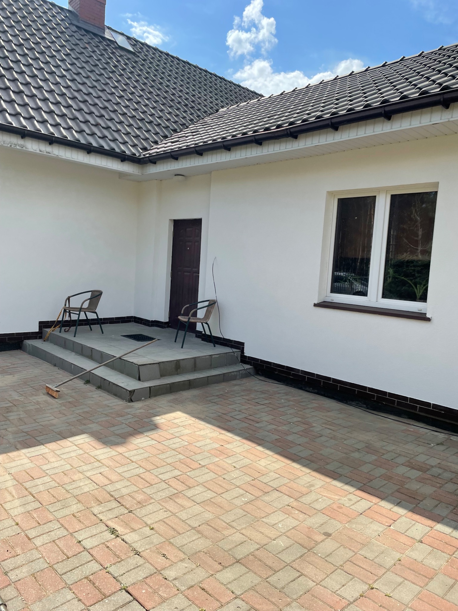 Dom z ciemnobrązowym dachem z dachówki ceramicznej, białymi ścianami, rynnami i brukowanym podjazdem. Na schodach prowadzących do drzwi wejściowych stoją dwa krzesła i leży miotła.