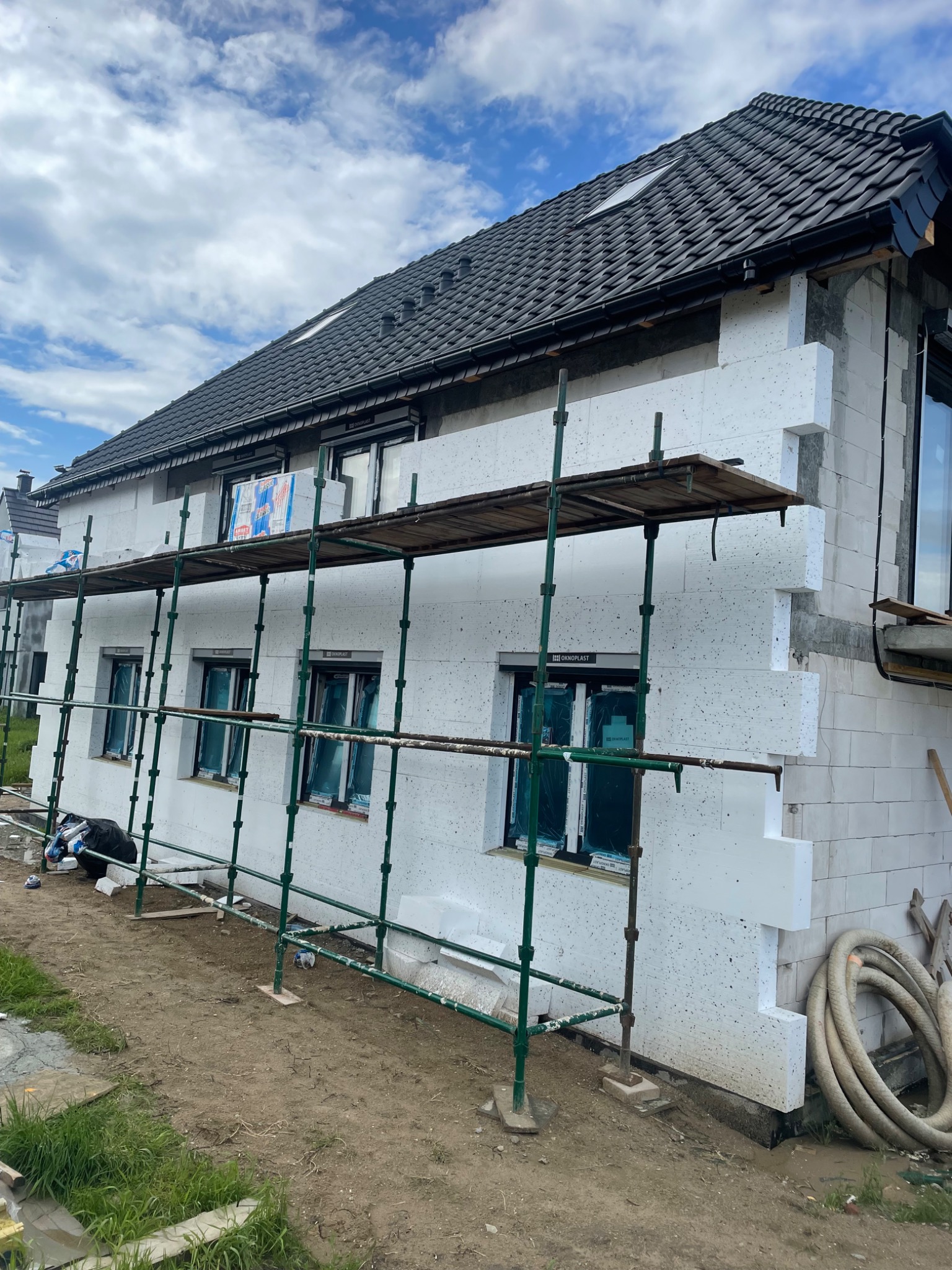 Dom w trakcie ocieplania, widoczne rusztowanie i płyty styropianowe na elewacji, dach pokryty czarną dachówką.