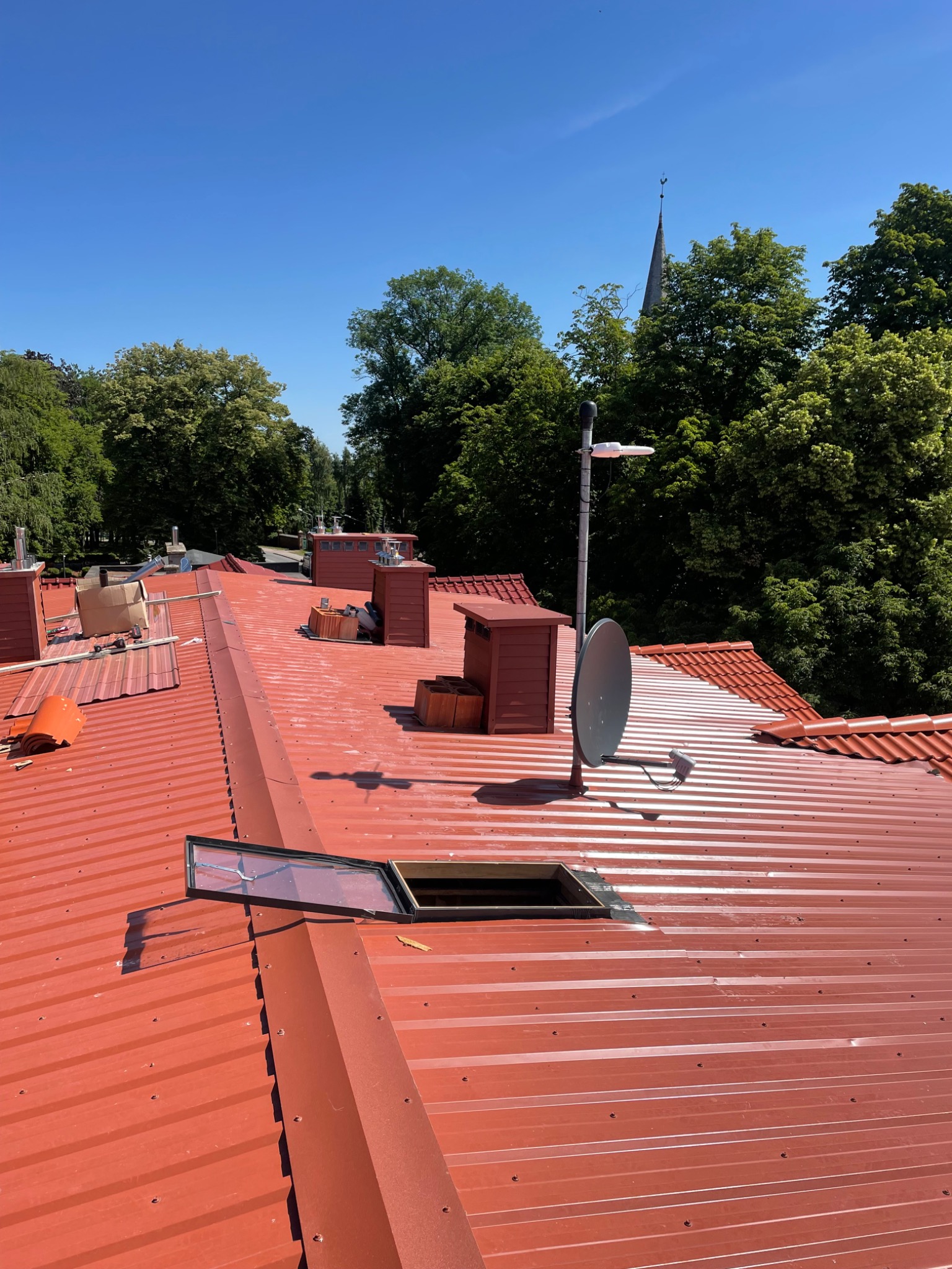 Czerwony blaszany dach z otwartym oknem dachowym i kominami na tle zielonych drzew i błękitnego nieba.