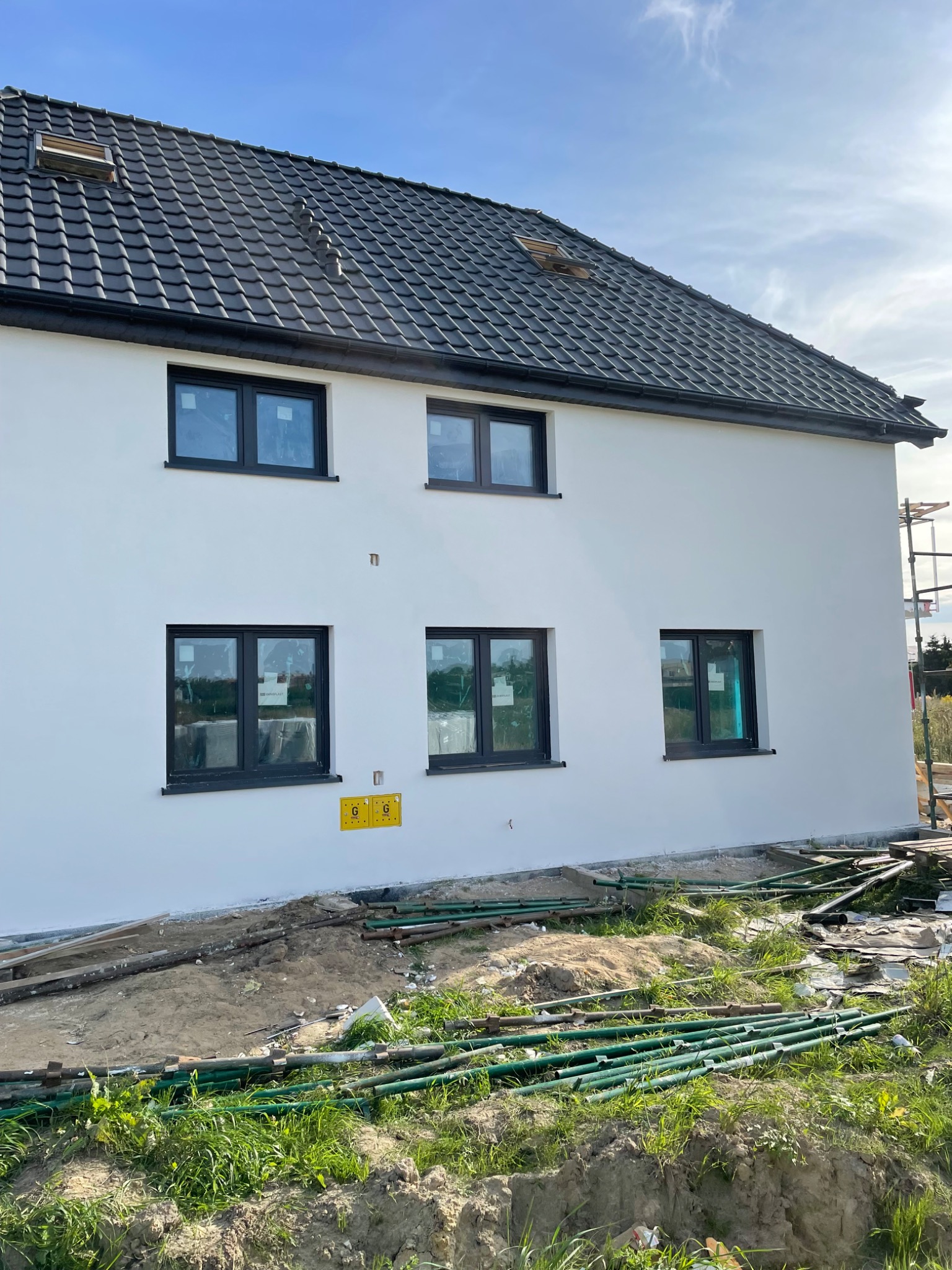 Nowy dom jednorodzinny z ciemną dachówką i czarnymi oknami, widok z boku na elewację w trakcie wykończenia, z widocznymi rurami i elementami rusztowania na ziemi.