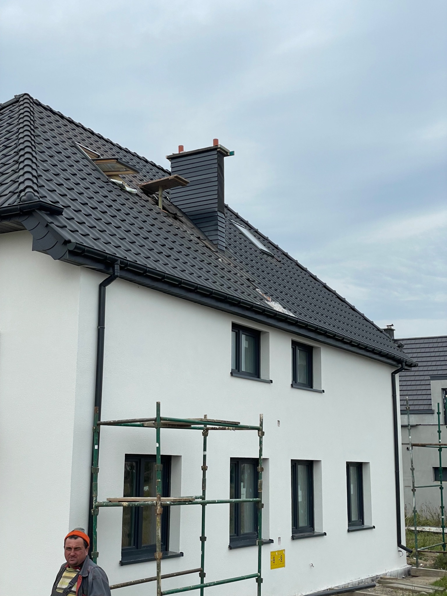 Nowy, ciemny dach z dachówki ceramicznej na białym domu, częściowo podparty rusztowaniem, widoczne okno dachowe i komin z elementami deskowania, pracownik w dolnym rogu kadru.