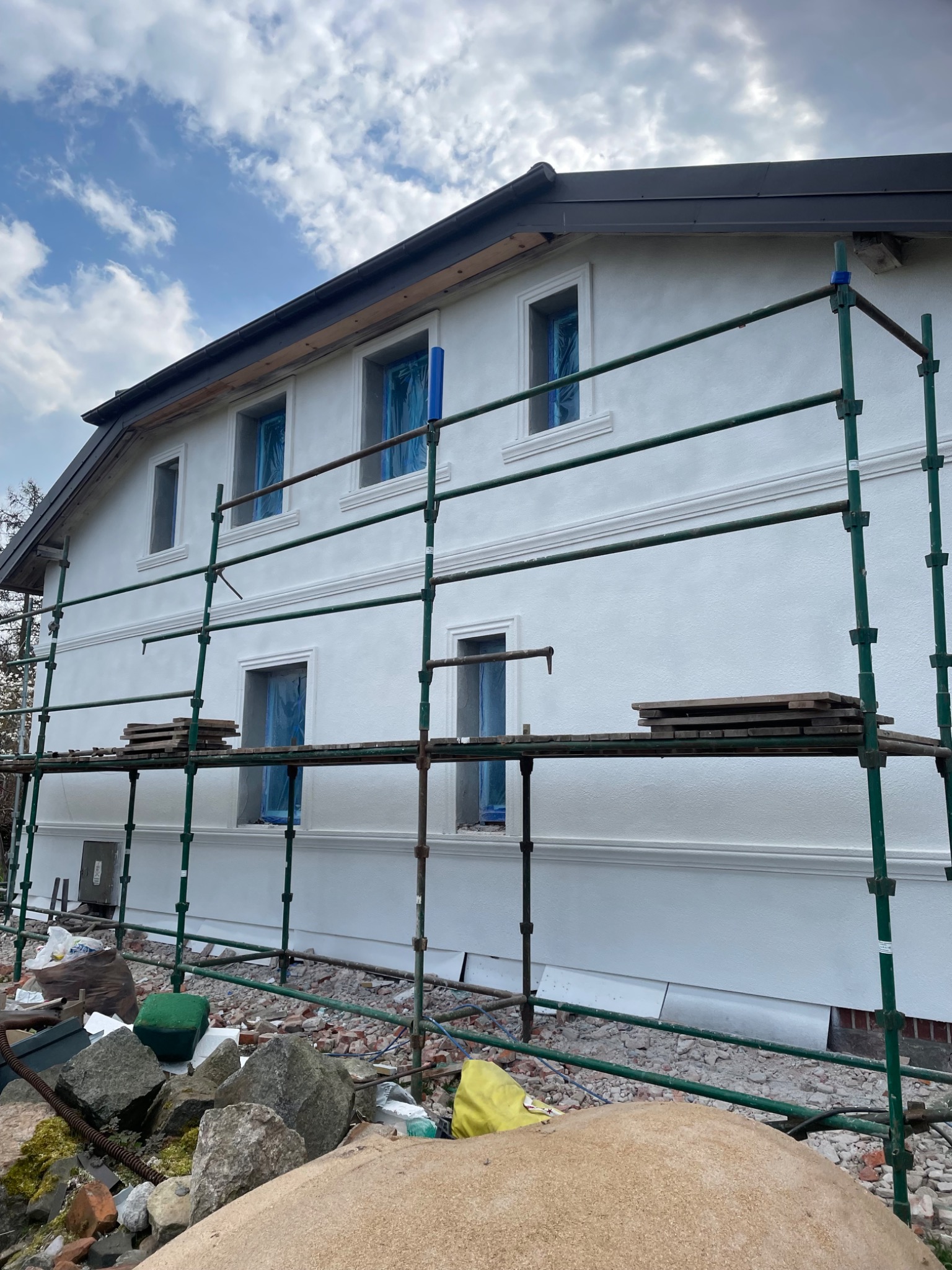 Dom jednorodzinny w trakcie prac elewacyjnych, z widocznym rusztowaniem, zabezpieczonymi oknami i nowym, ciemnym wykończeniem okapu.
