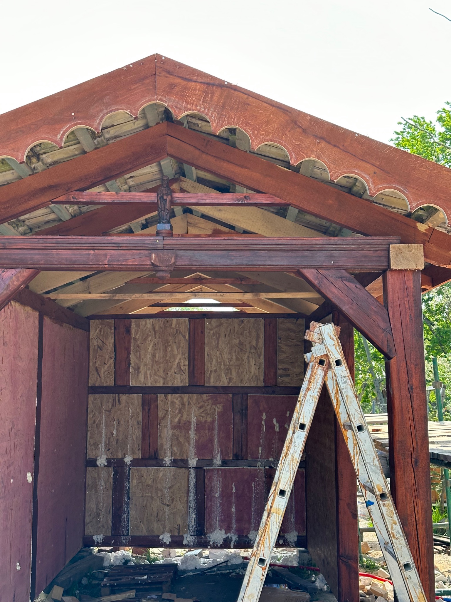 Konstrukcja altany z drewnianą więźbą dachową i dekoracyjnym wykończeniem okapu, widoczna drabina oparta o filar.