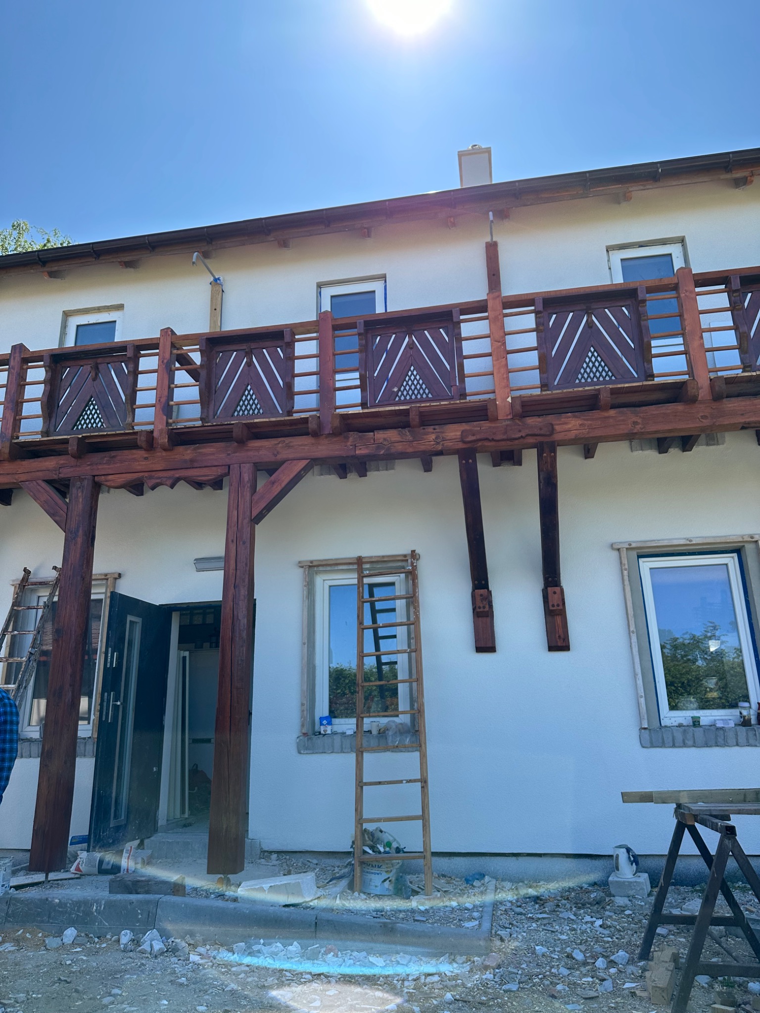 Drewniany balkon z ozdobnymi panelami w trakcie budowy, drabiny opierają się o ścianę, widoczne materiały budowlane i narzędzia na zewnątrz budynku.