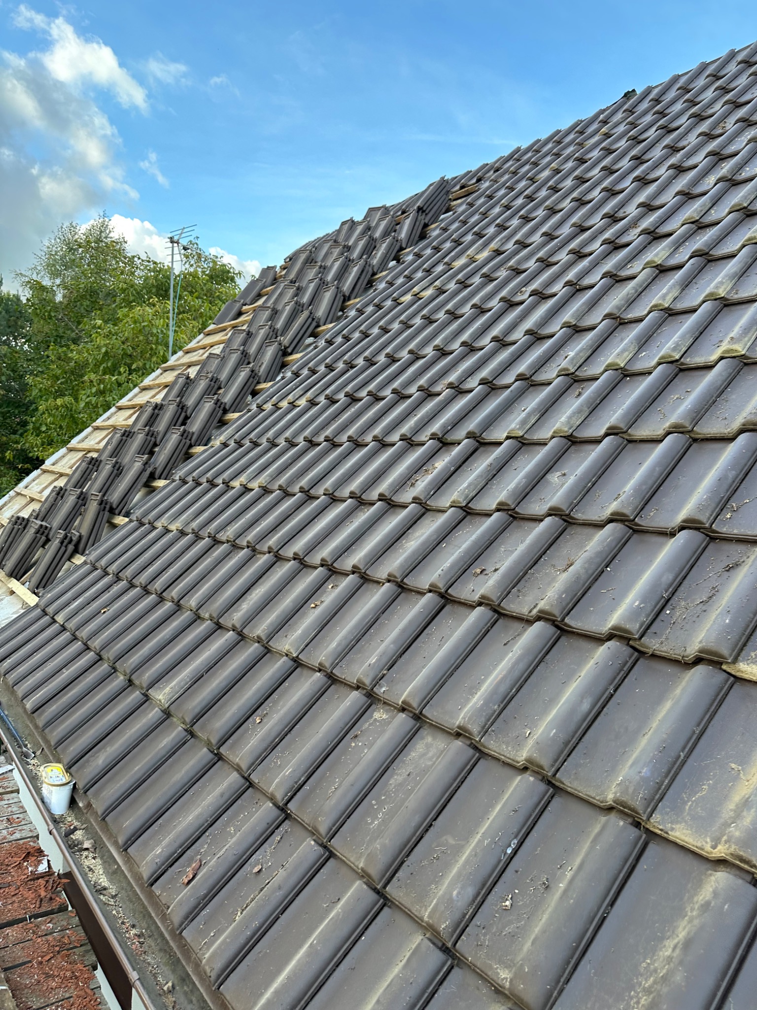 Częściowo ułożone, ciemnobrązowe dachówki ceramiczne na drewnianej konstrukcji dachu, widoczne drzewa i błękitne niebo z chmurami w tle.