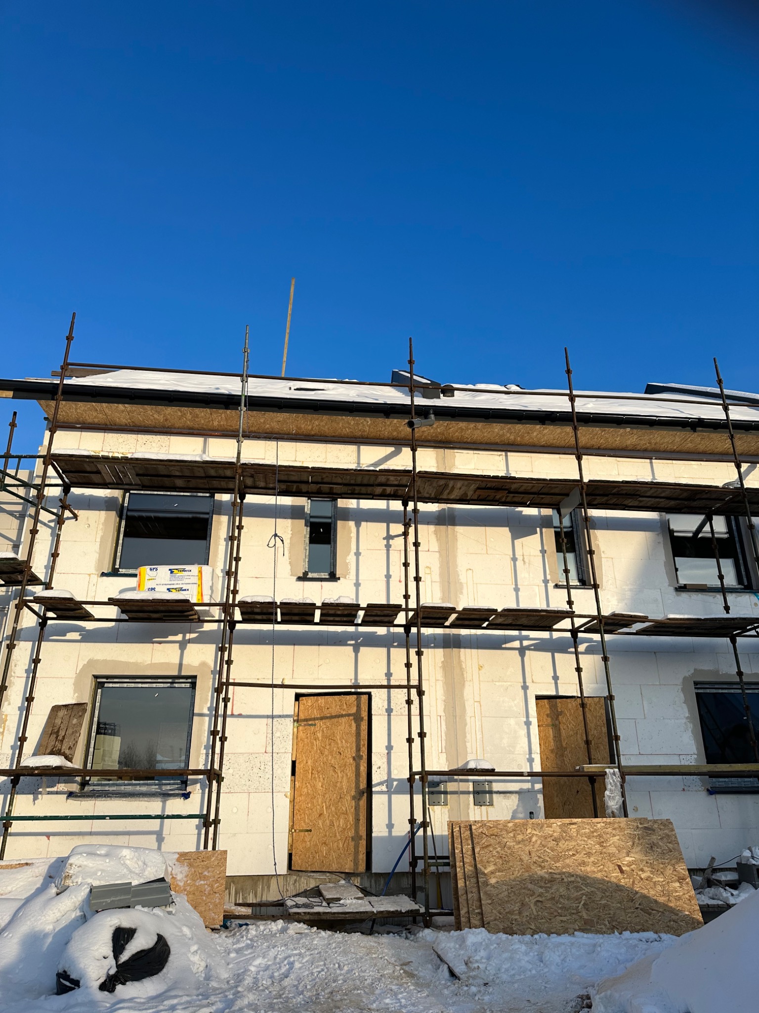 Budynek w trakcie budowy z białych bloków, otoczony rusztowaniami, z tymczasowymi drzwiami z płyty OSB i oknami, widoczny materiał izolacyjny na rusztowaniu, śnieg na ziemi i częściowo na dachu...