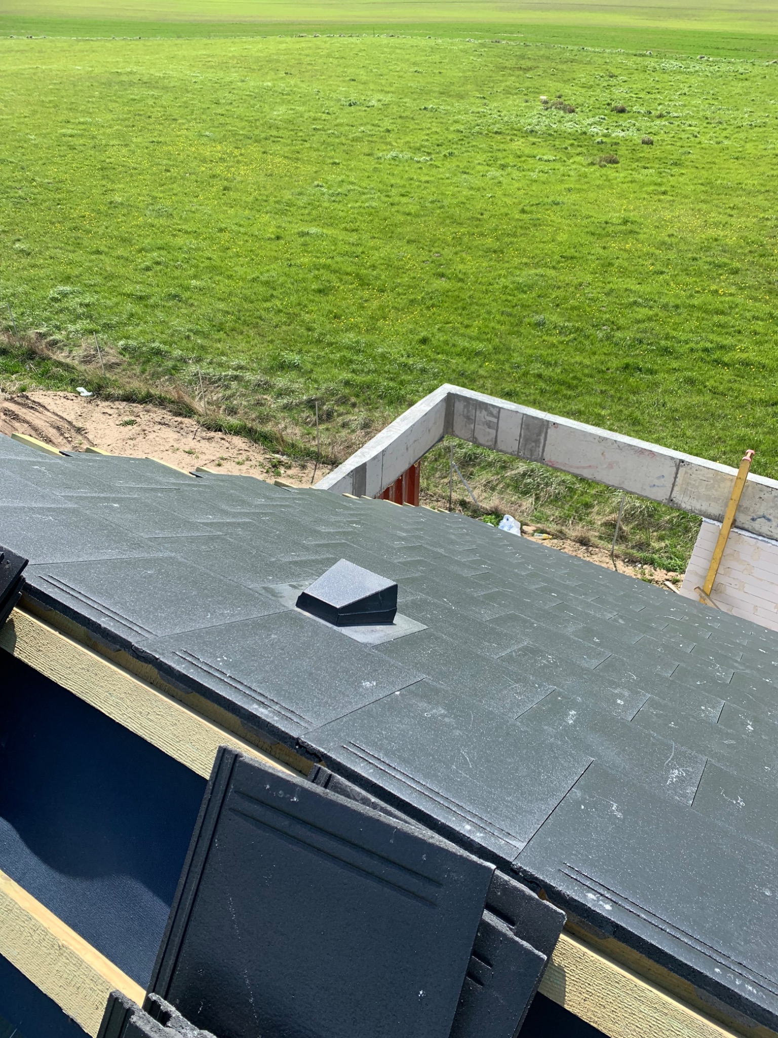 Montaż czarnej, matowej dachówki na membranie na tle zielonego pola i częściowo ukończonej konstrukcji budynku.