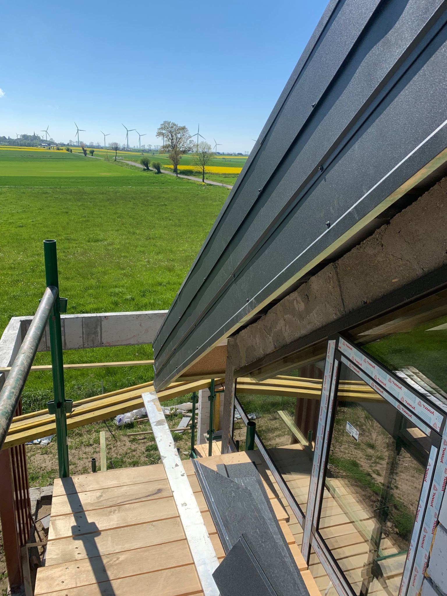 Montaż blachy na dachu budynku w trakcie budowy, widoczne rusztowania, okna i krajobraz z polami i wiatrakami w tle.