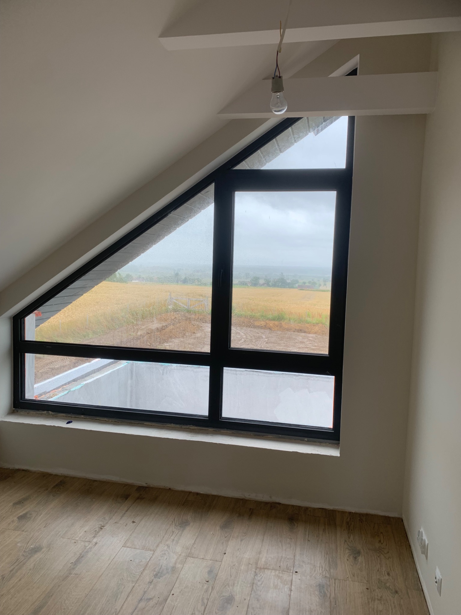 Nowe okno dachowe z czarną ramą w Jeżyczkach, widok na pole w pochmurny dzień, surowy stan wykończenia wnętrza.