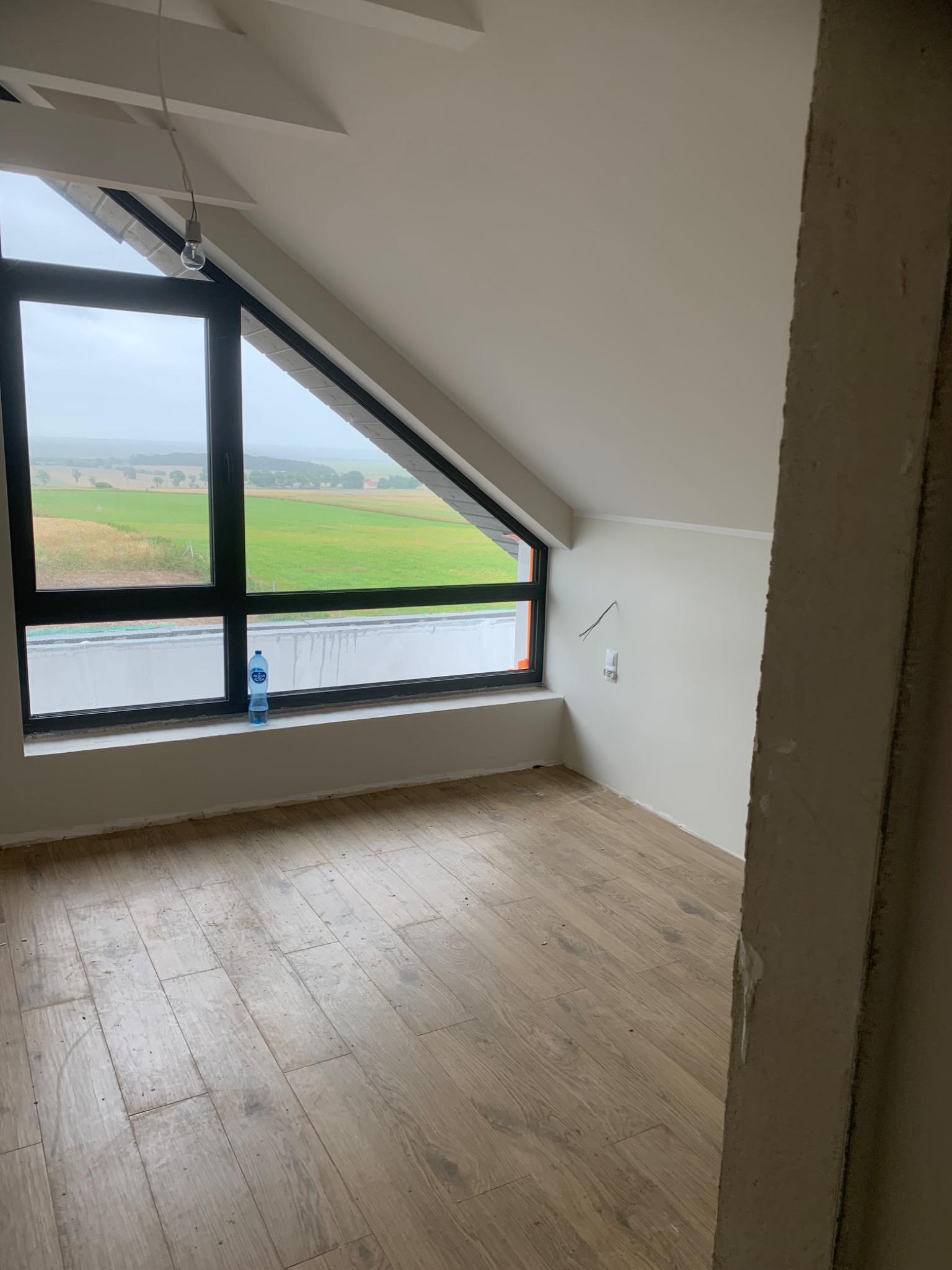 Poddasze w trakcie wykańczania z oknem dachowym i widokiem na zielone pola, butelka wody na parapecie, surowy betonowy filar w pierwszym planie.
