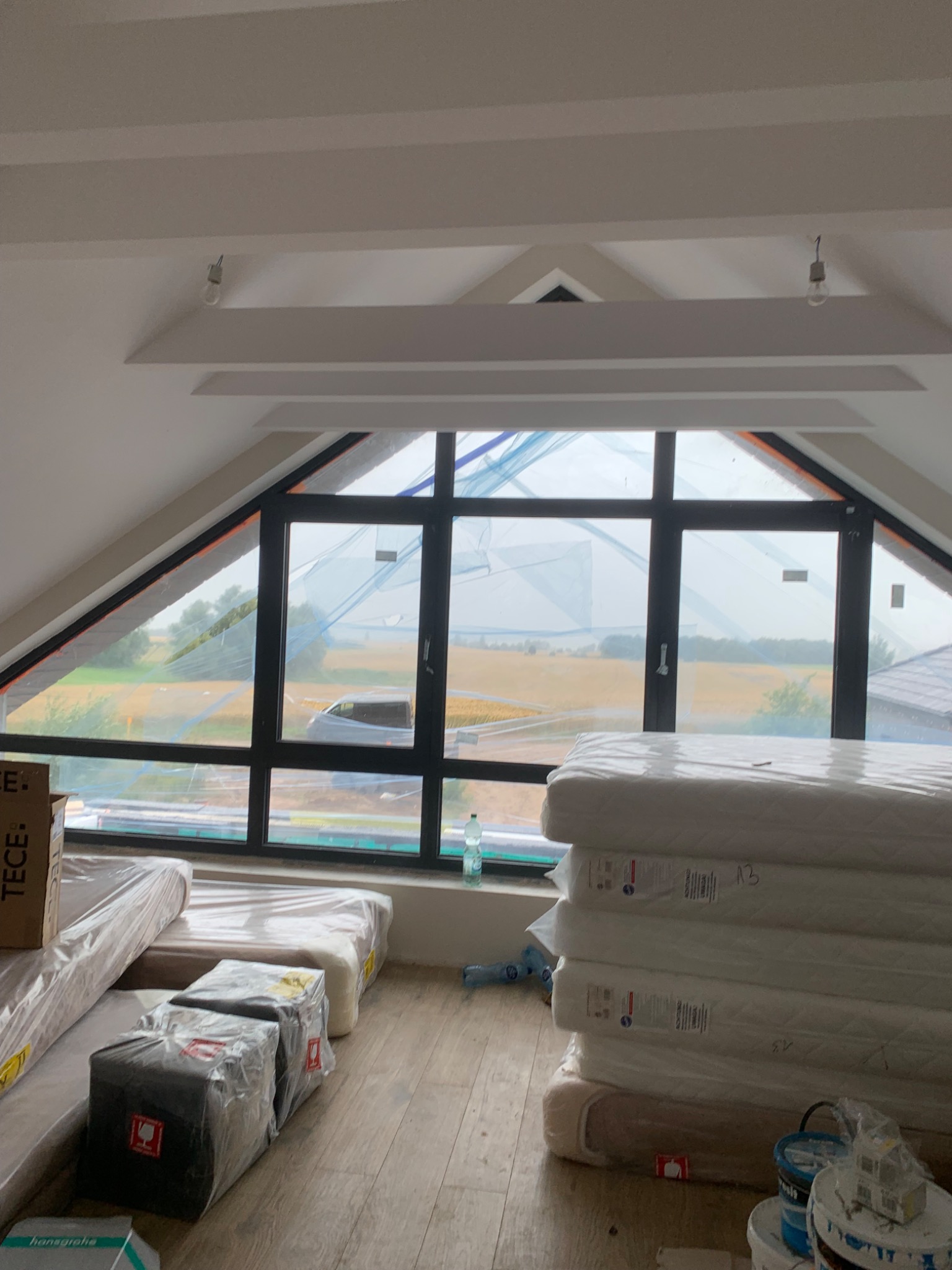 Poddasze w trakcie prac wykończeniowych, widoczne okno dachowe z widokiem na pole, materiały budowlane i pakiety z nowymi materacami.