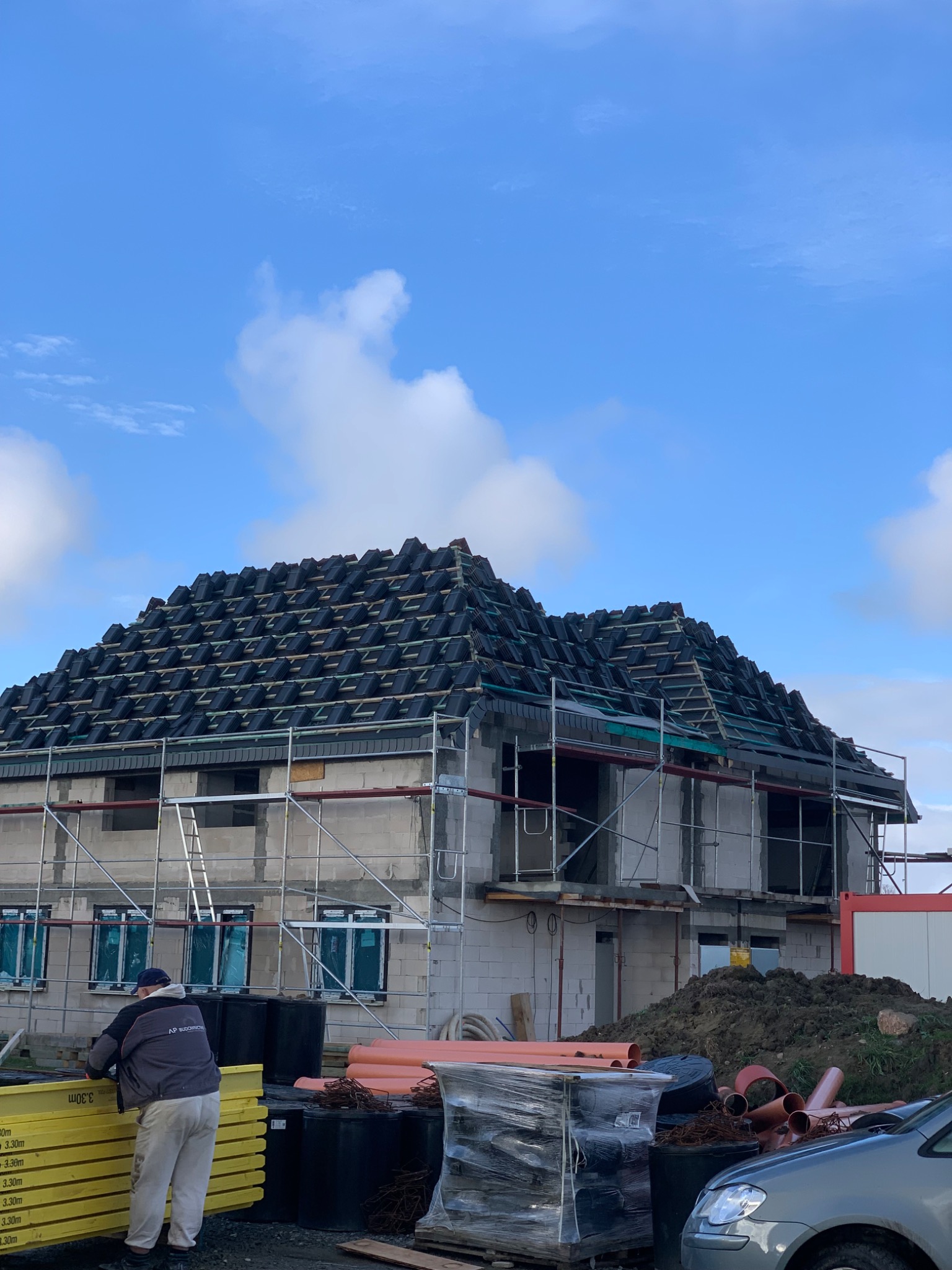 Dom w trakcie budowy z częściowo położonym czarnym dachem, widoczne rusztowania, okna zabezpieczone folią, oraz stos żółtych desek i pomarańczowych rur na pierwszym planie.