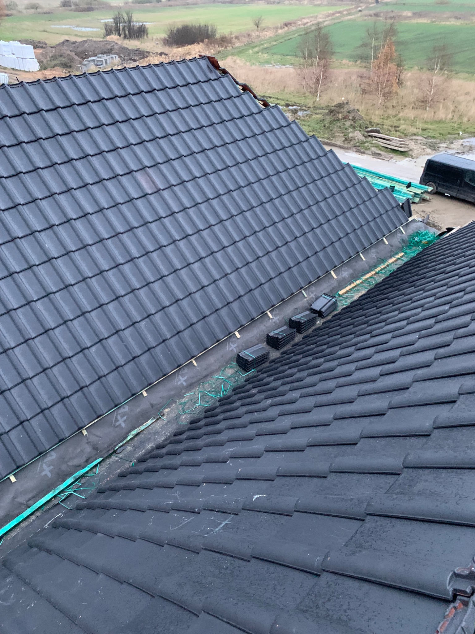 Układanie ciemnej dachówki na dachu domu, widoczna membrana dachowa i zabezpieczenia krawędzi, w tle krajobraz z polami i drzewami.