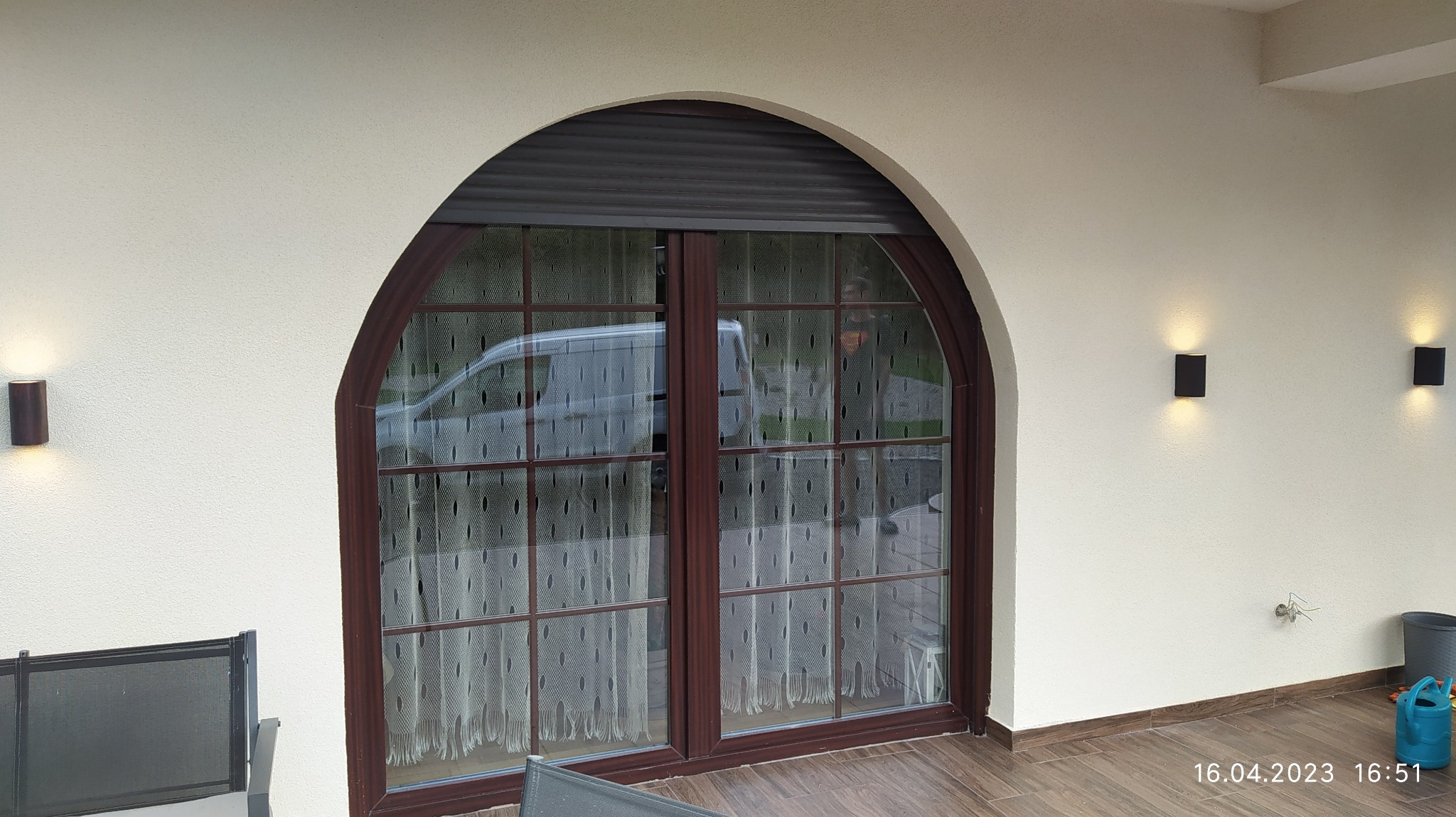 Zamknięte dwuskrzydłowe okno łukowe z brązową ramą i roletą zewnętrzną, oświetlone czarnymi kinkietami na elewacji.