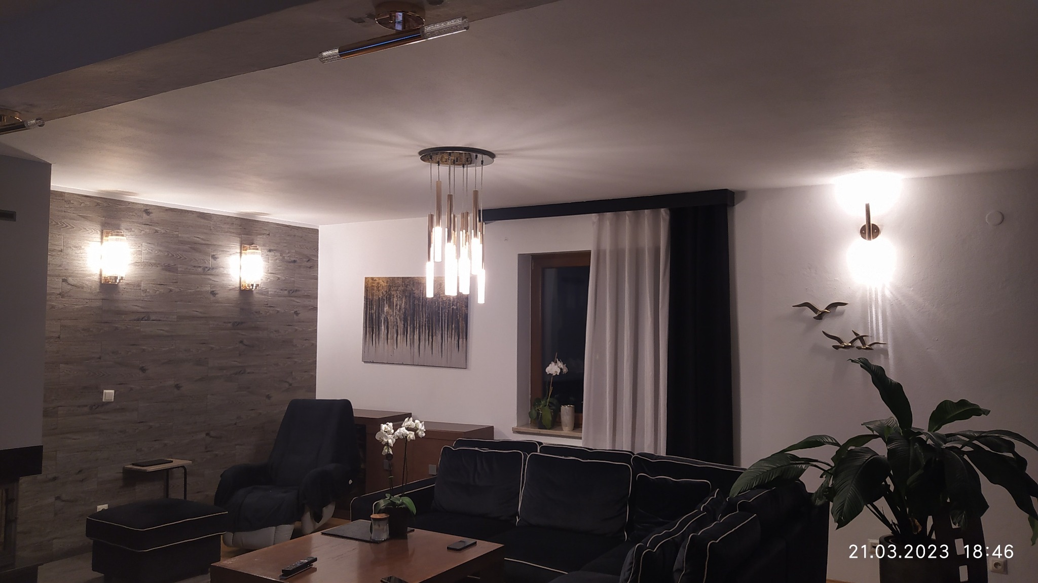 Salon z trzema rodzajami oświetlenia: wisząca lampa z podłużnymi kloszami, kinkiety na drewnianej ścianie oraz pojedynczy kinkiet ścienny z dekoracją w postaci ptaków.