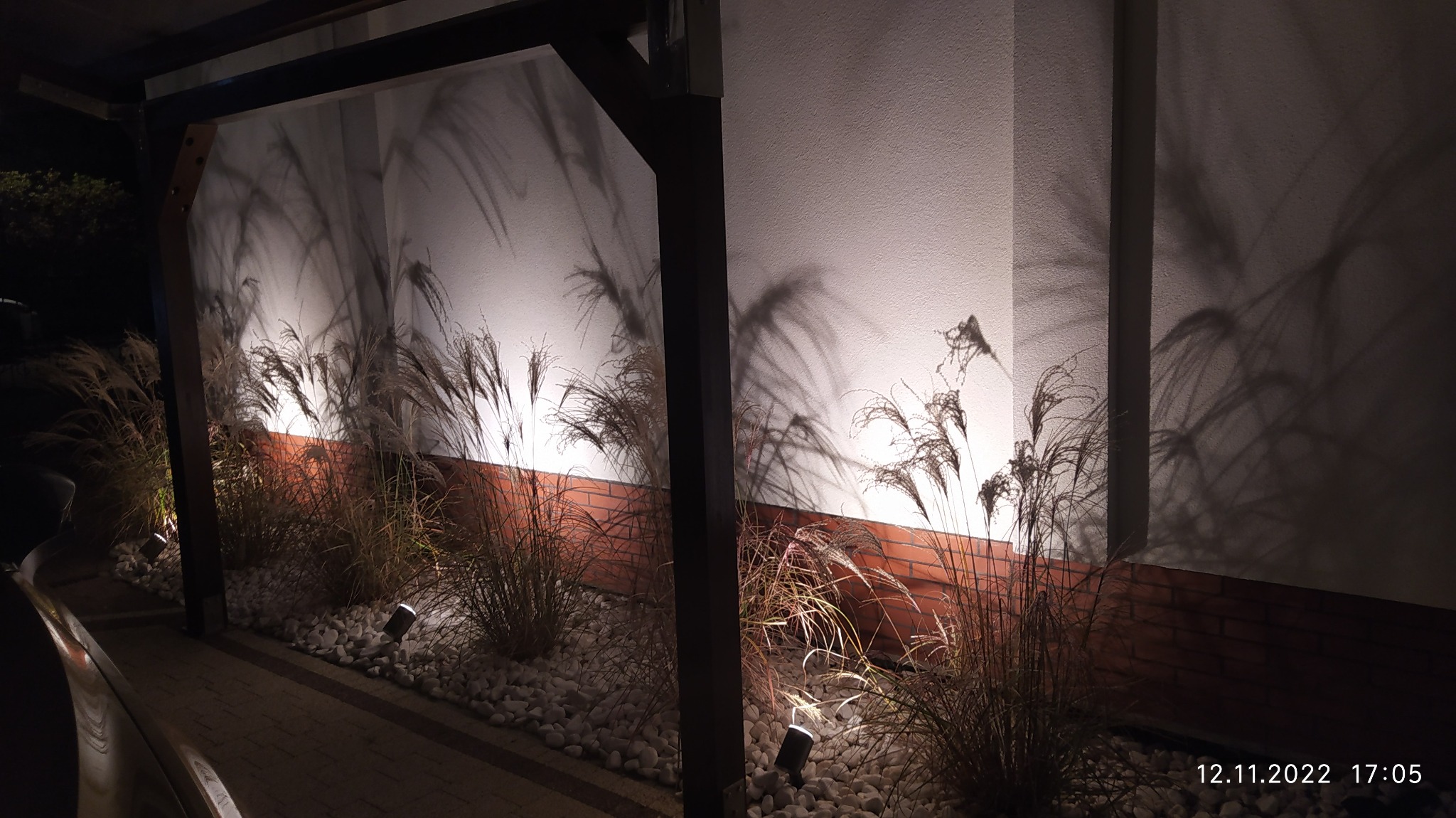 Oświetlenie dekoracyjne ogrodu nocą: lampy gruntowe oświetlają ozdobne trawy, rzucając cienie na białą ścianę i ceglany murek, z elementami drewnianej konstrukcji w tle.