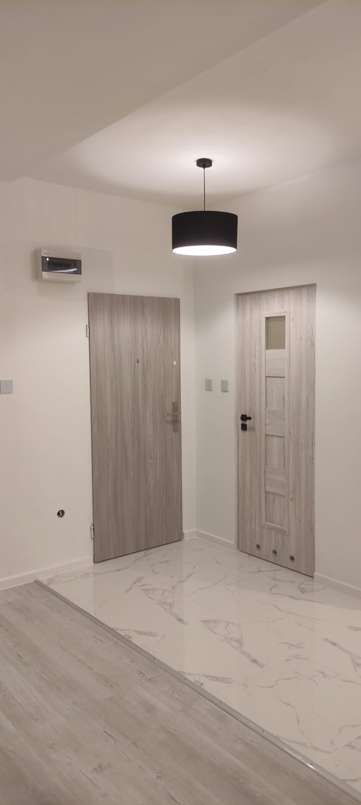 Wnętrze po remoncie: dwie drewnopodobne drzwi, czarna lampa wisząca z abażurem, białe ściany i marmurowe płytki podłogowe, widoczna skrzynka elektryczna na ścianie.