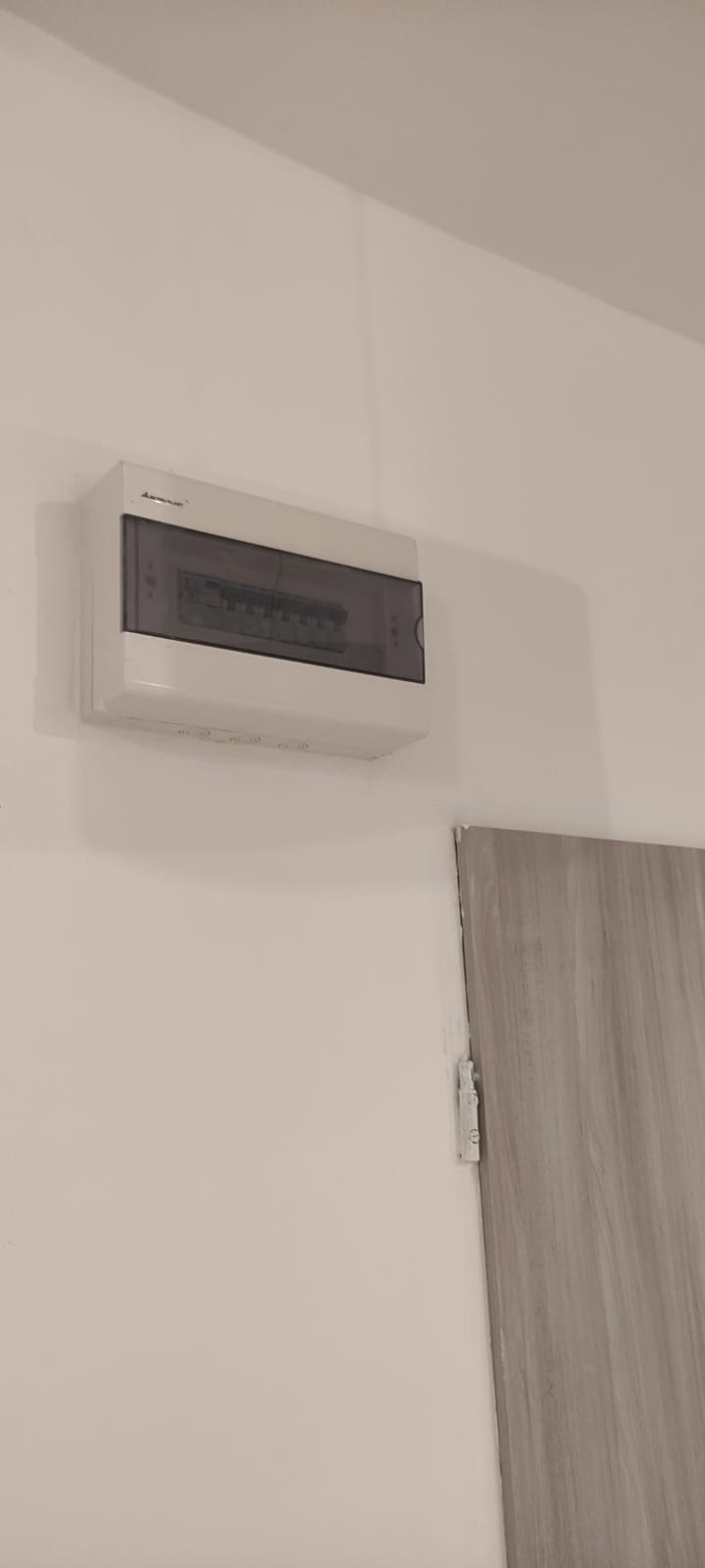 Zamknięta, biała skrzynka rozdzielcza z szarą, przezroczystą pokrywą zamontowana na białej ścianie, obok fragmentu szarej ściany lub drzwi.