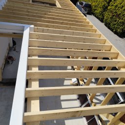 STUDZIŃSKI KONSTRUKCJE Szymon Studziński - Konstrukcja drewniana z belek, prawdopodobnie zadaszenia tarasu, w trakcie budowy. Widoczne elementy mocujące i narzędzia w tle. Jasne drewno, dach z dachówki w tle.