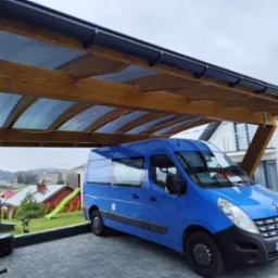 Niebieski bus pod drewnianą wiatą z przezroczystym zadaszeniem, na posesji z kostki brukowej. W tle dom i zieleń.