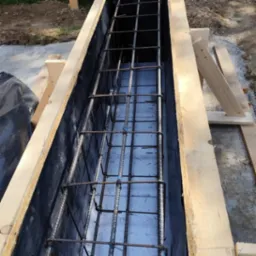 Szczegółowe ujęcie z góry na szalunek zbrojeniowy z metalowymi prętami, przygotowany do zalania betonem. Widoczne deski szalunkowe i folia ochronna.