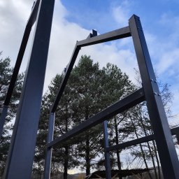 Metalowa konstrukcja szkieletowa w trakcie montażu, widok z dołu na tle drzew i błękitnego nieba z chmurami. Elementy skręcane śrubami.