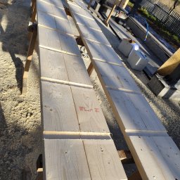 Długie, jasne deski na drewnianych kozłach, przygotowywane do montażu. W tle narzędzia i elementy konstrukcyjne. Widoczne ślady obróbki drewna.