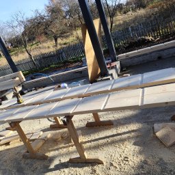 Montaż drewnianych elementów konstrukcyjnych na placu budowy. Widoczne narzędzia i drewniane podpory. W tle ogrodzenie i zieleń.