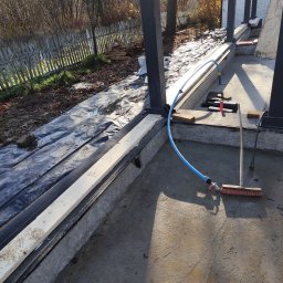 Wylewka fundamentowa z hydroizolacją i deskowaniem, przygotowana pod budowę. Widoczny wąż z wodą i narzędzia budowlane. W tle ogrodzenie i drzewa.