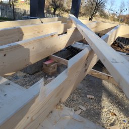 STUDZIŃSKI KONSTRUKCJE Szymon Studziński - Konstrukcja szkieletowa z jasnego drewna w trakcie montażu, widoczne elementy fundamentu i słupy nośne. W tle drzewa i ogrodzenie.