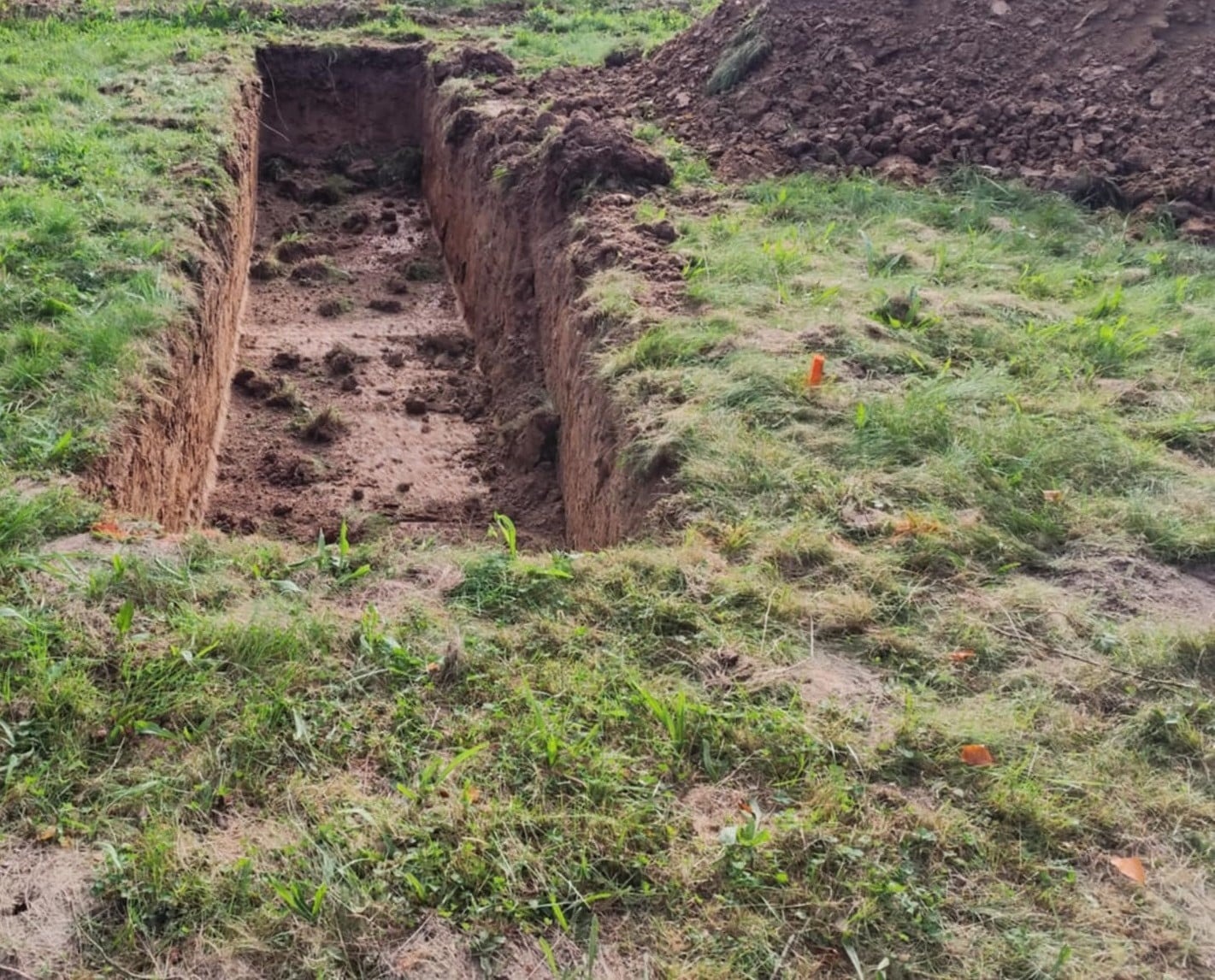 Wykop pod fundament z widocznymi ścianami wykopu i stertą ziemi w tle, otoczony trawą. W ziemi wbite pomarańczowe znaczniki.