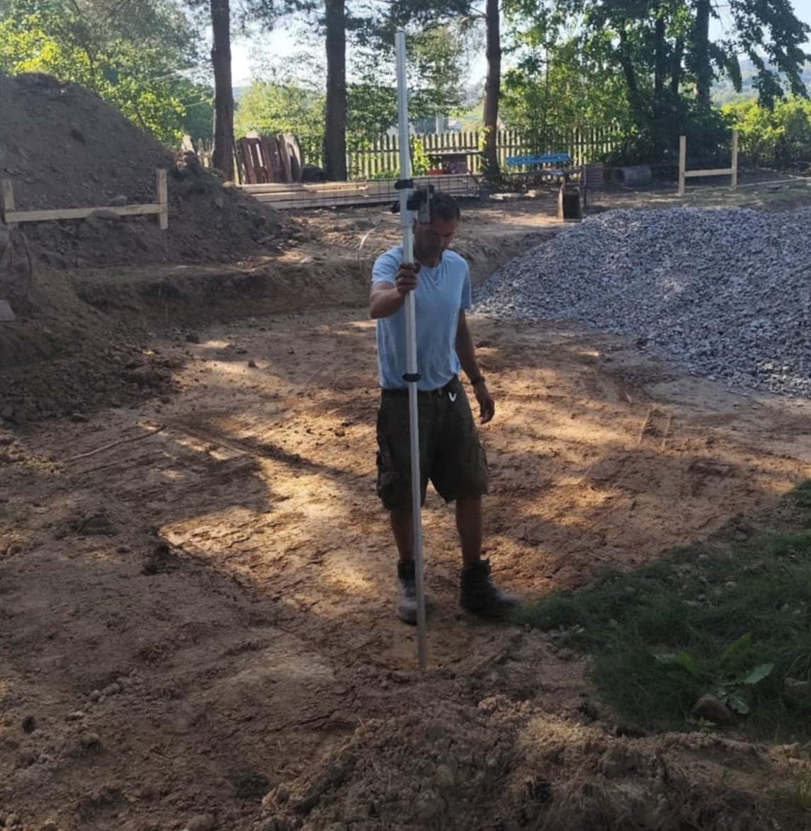 Pracownik budowlany mierzy poziom gruntu niwelatorem optycznym na wykopie pod fundamenty domu. W tle hałda ziemi i kamieni.