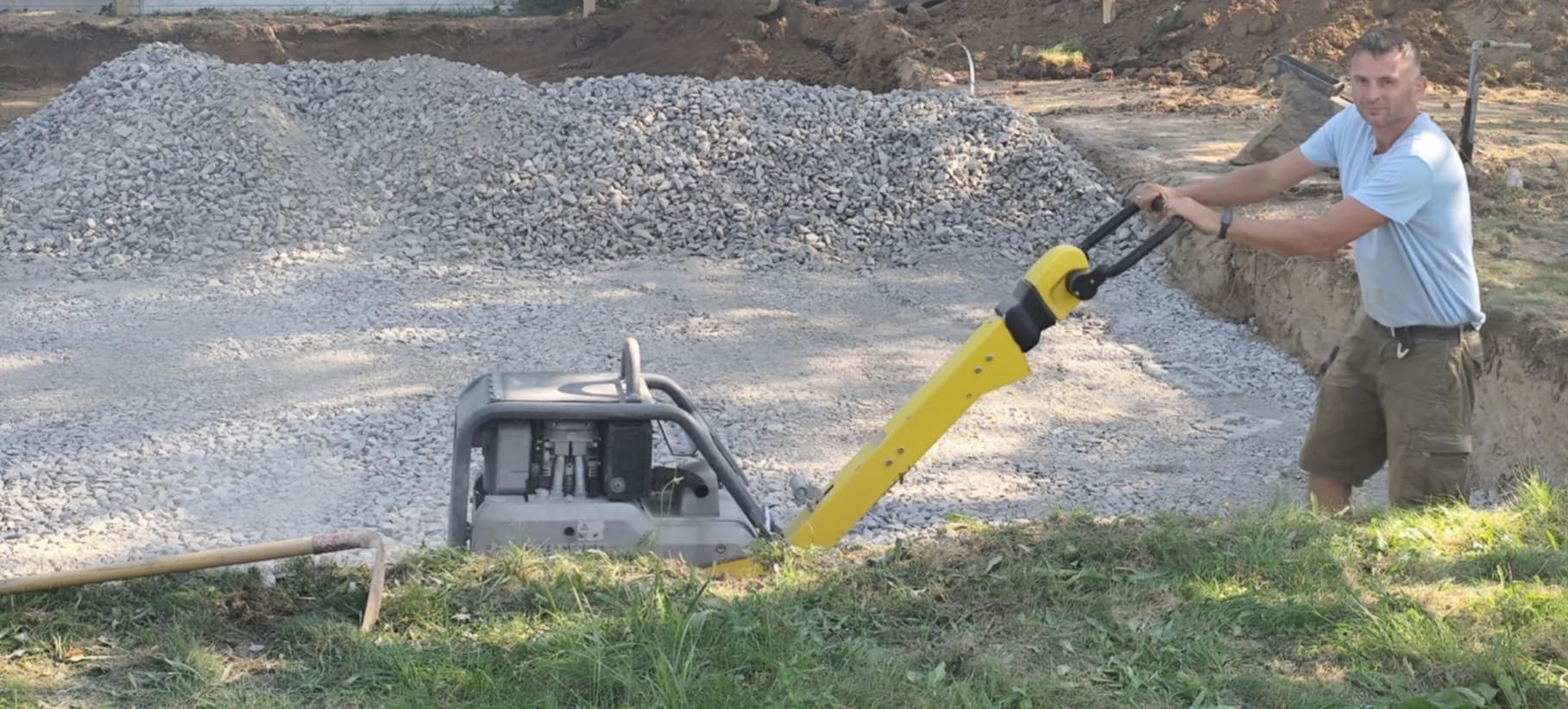Pracownik zagęszcza podłoże pod fundamenty ubijarką w wykopie, obok sterta gruzu i narzędzie. Prace ziemne w toku, przygotowanie terenu pod budowę.