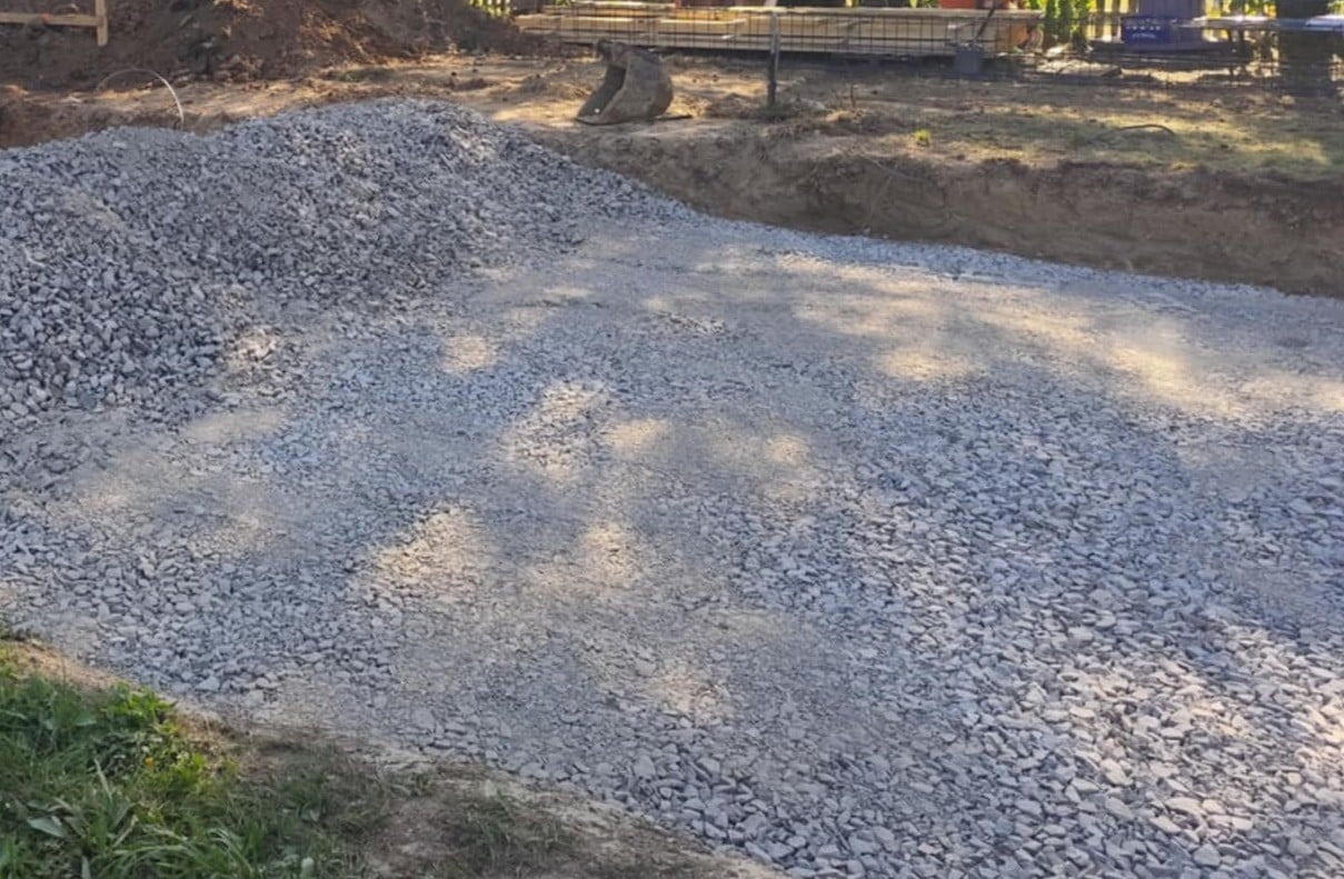Przygotowany teren pod budowę: wyrównany grunt z nasypem z szarego kruszywa, widoczne wykopy ziemne i elementy konstrukcji w tle.
