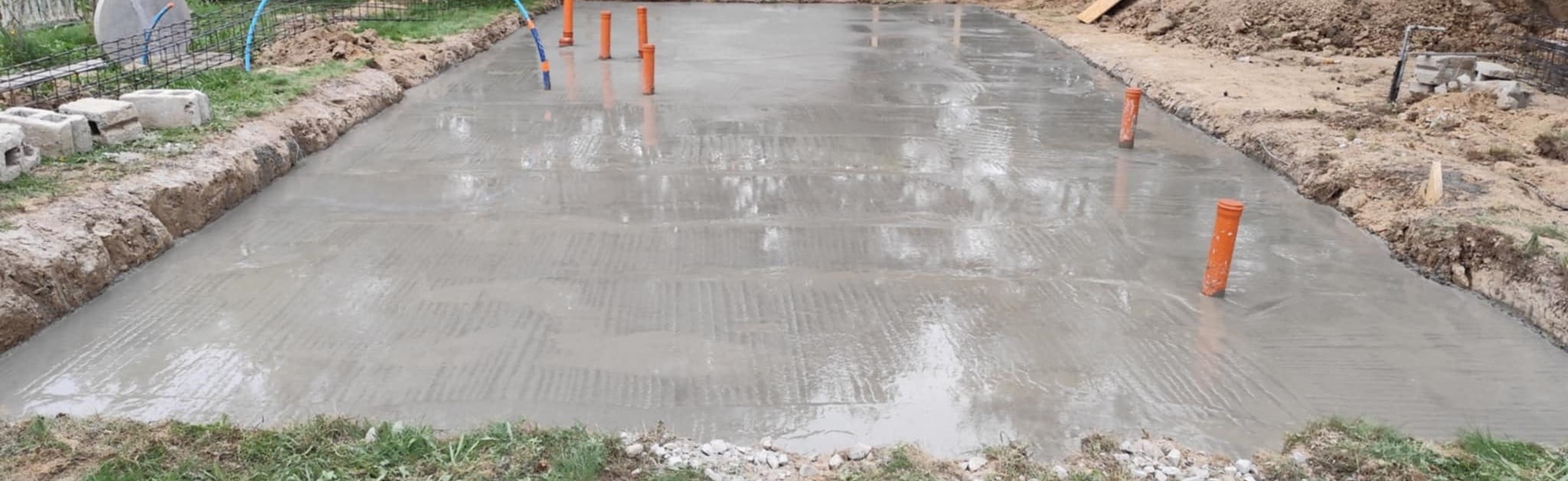 Świeżo wylany fundament z wystającymi pomarańczowymi rurami, widoczne ślady po szalunku. Teren wyrównany, przygotowany do dalszych prac budowlanych.