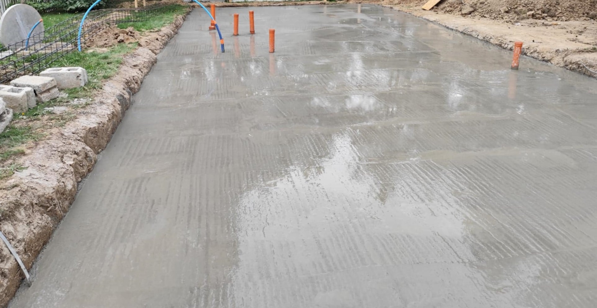 Świeżo wylany fundament pod budowę. Widoczne pionowe rury instalacyjne, obrzeża wykopu i zbrojenie. Jasne, naturalne oświetlenie.