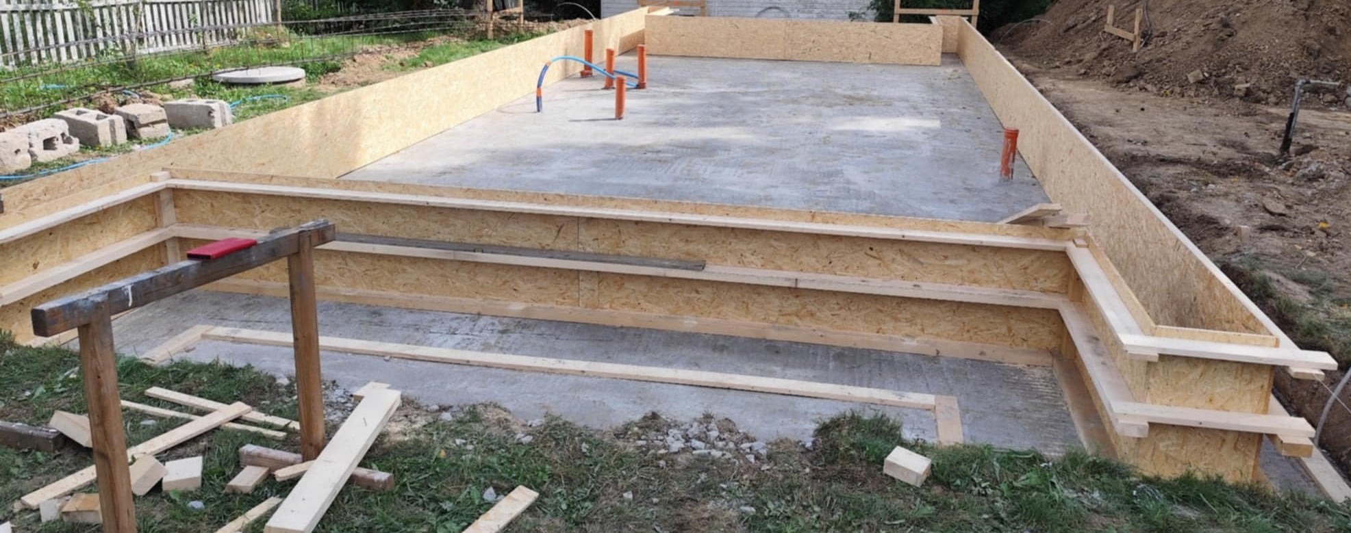 Deskowanie fundamentu domu jednorodzinnego z widocznymi pionowymi rurami instalacyjnymi i deskami do podtrzymania konstrukcji. Częściowo zalany beton.