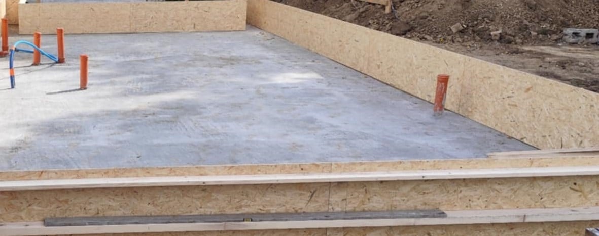 Zalany betonowy fundament z deskowaniem z płyty OSB i wystającymi rurami kanalizacyjnymi. Widoczny fragment terenu.