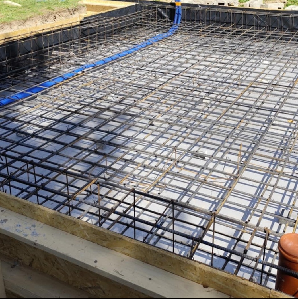 Zbrojenie fundamentu z widoczną niebieską rurą instalacyjną, przygotowane do zalania betonem. Szalunek w tle. Widok z góry na siatkę z prętów.