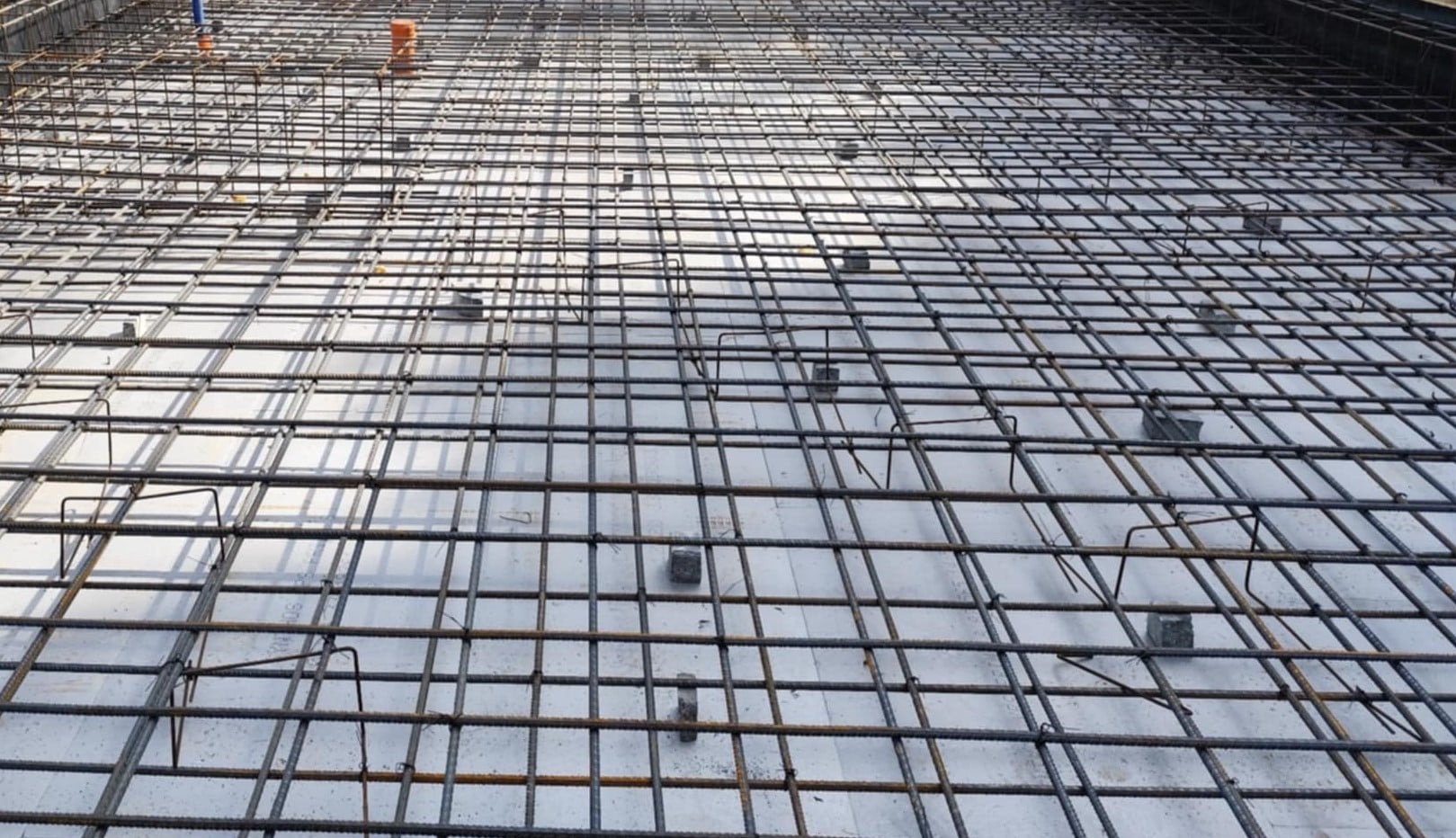 Zbrojenie płyty fundamentowej na białej izolacji, widok z góry. Stalowe pręty tworzą gęstą siatkę. Elementy dystansowe podtrzymują zbrojenie.