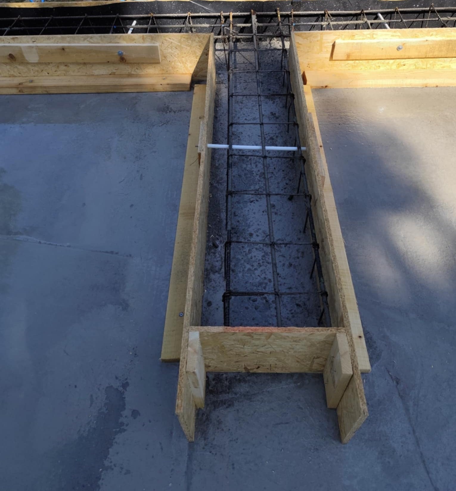 Szczegółowe ujęcie z góry na zbrojenie fundamentu w drewnianym szalunku, z widoczną stalową siatką i elementami konstrukcyjnymi na betonowej płycie.