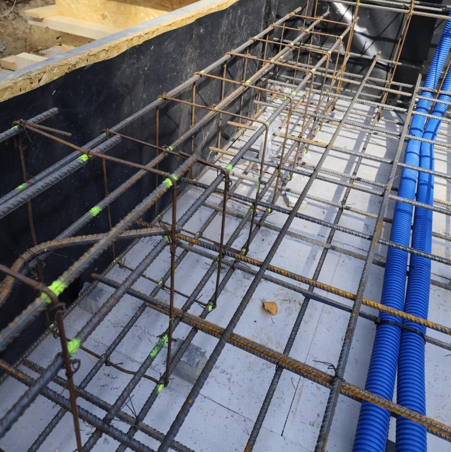 Zbrojenie fundamentu z widocznymi prętami i niebieskimi rurami w szalunku. Elementy konstrukcyjne przygotowane do zalania betonem.